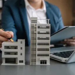 Mężczyzna w garniturze analizuje modele budynków, trzymając długopis nad modelem niskiego budynku, obok kalkulator i laptop