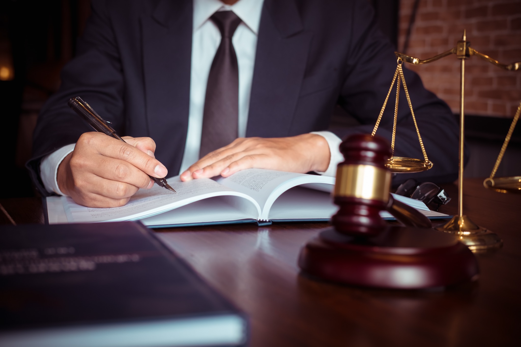 Mężczyzna w garniturze podpisuje dokument piórem, obok waga sprawiedliwości i młotek sędziowski.