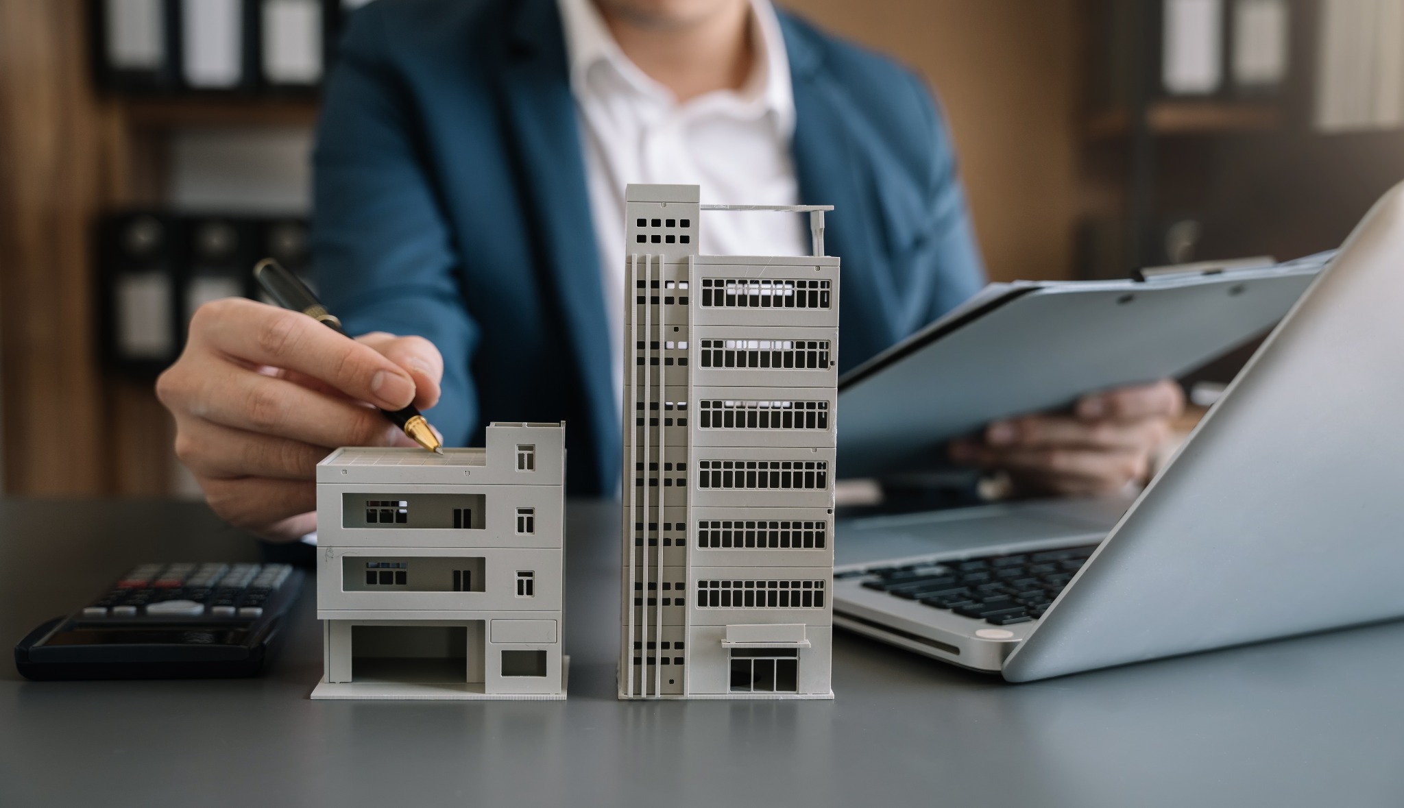 Mężczyzna w garniturze analizuje modele budynków, trzymając długopis nad modelem niskiego budynku, obok kalkulator i laptop