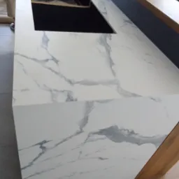 Biały blat z marmurowym wzorem, wycięty otwór na zlew, widoczny fragment nogi osoby w tle, montaż blatu w trakcie.