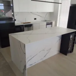 Nowoczesna kuchnia z wyspą, białe szafki, blat kamienny z marmurkowym wzorem, czarna lodówka i regał na wino wbudowany w wyspę, jasna drewniana podłoga.