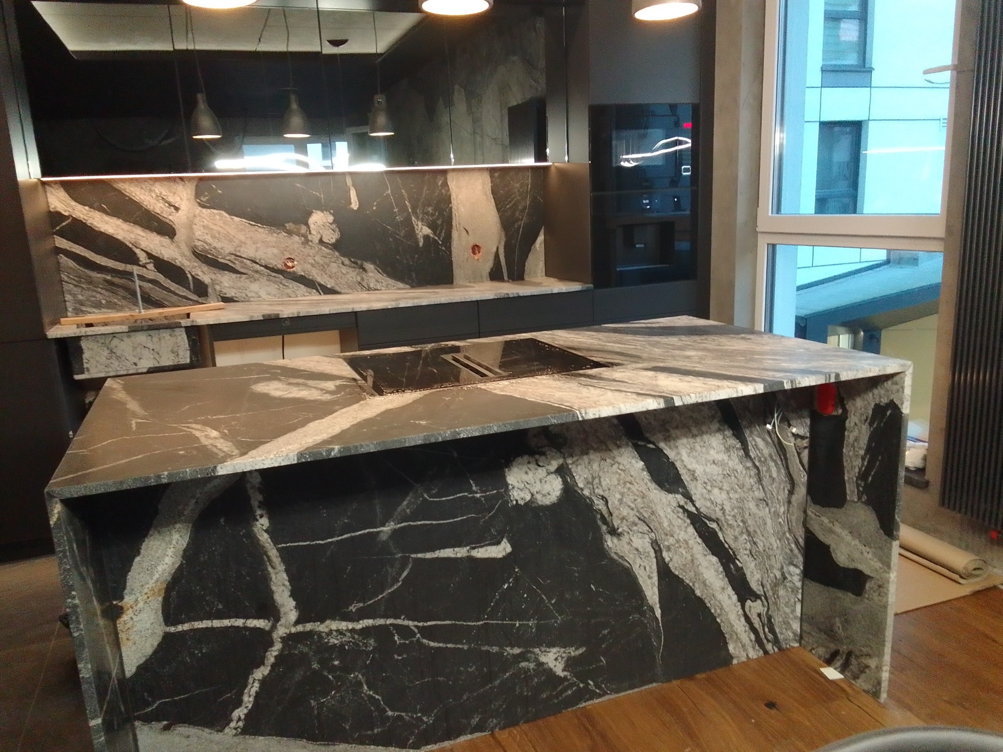 Nowoczesna kuchnia z wyspą pokrytą czarnym kamieniem z białymi żyłami, widoczne oświetlenie punktowe i zabudowane sprzęty AGD.