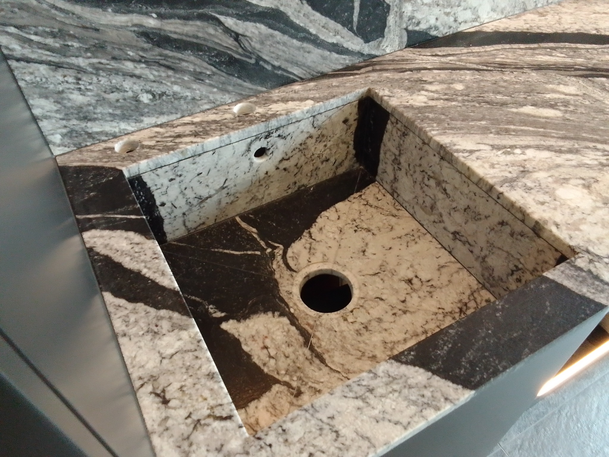 Wycięty otwór w blacie z szaro-białego kamienia, prawdopodobnie pod zlew, widoczne otwory na baterię i dozownik mydła, tło z kamienia o podobnym wzorze.