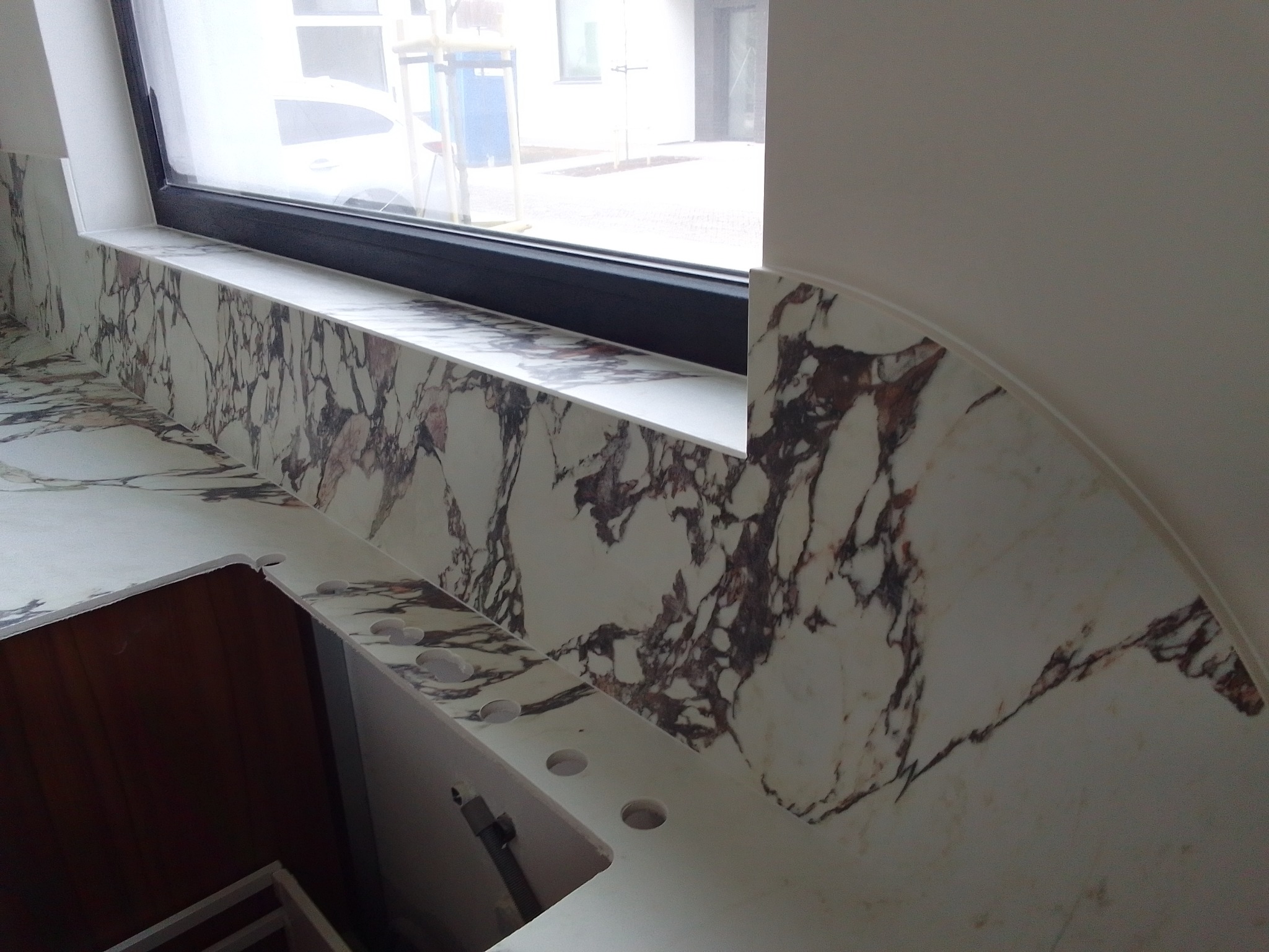 Fragment kuchennego blatu z białego marmuru z brązowymi żyłami, zamontowanego przy oknie, widoczne otwory na armaturę.
