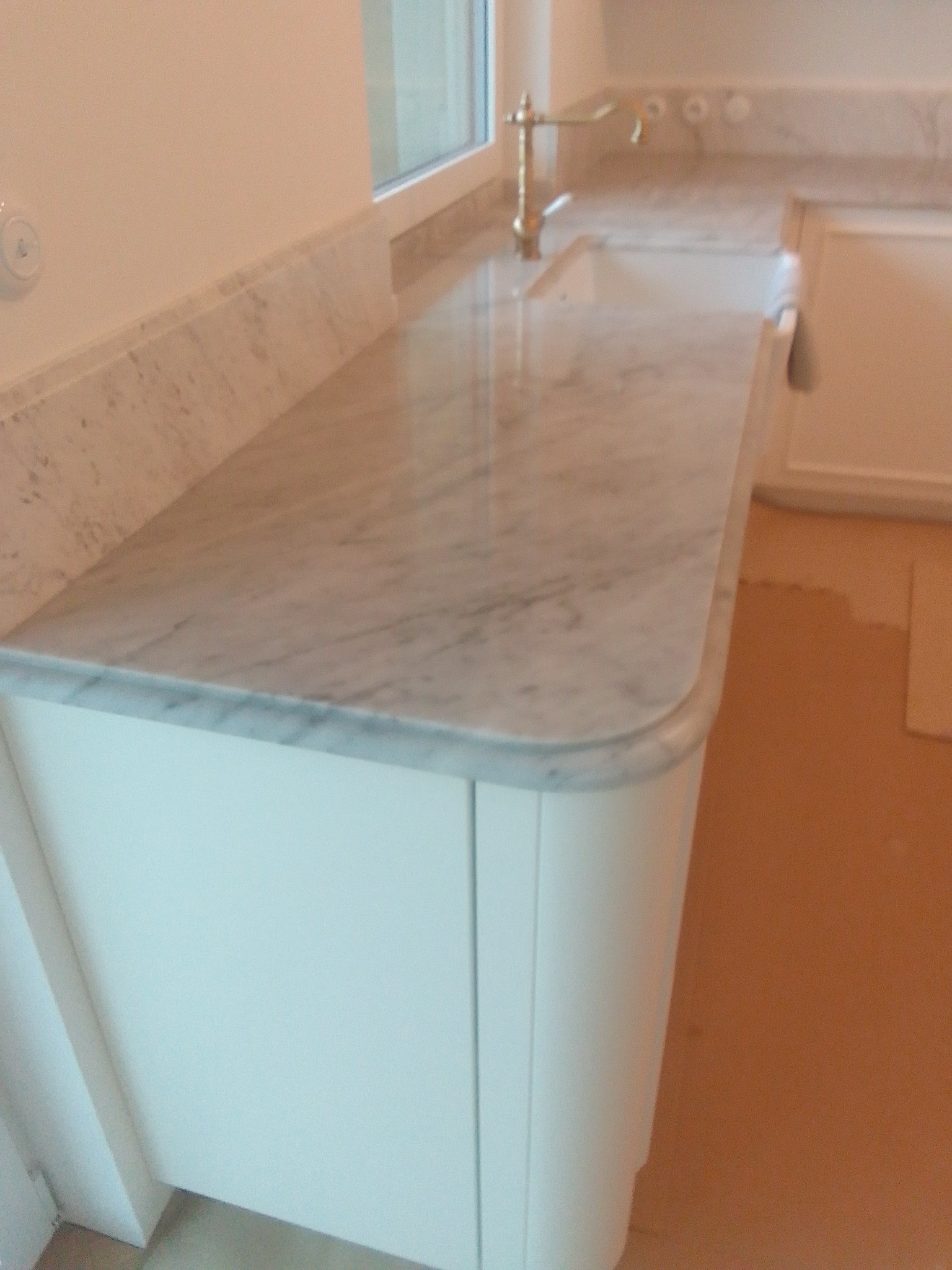 Jasny marmurowy blat z zaokrąglonym brzegiem, zamontowany na białych szafkach kuchennych, widoczna złota bateria umywalkowa i fragment okna z naturalnym światłem.