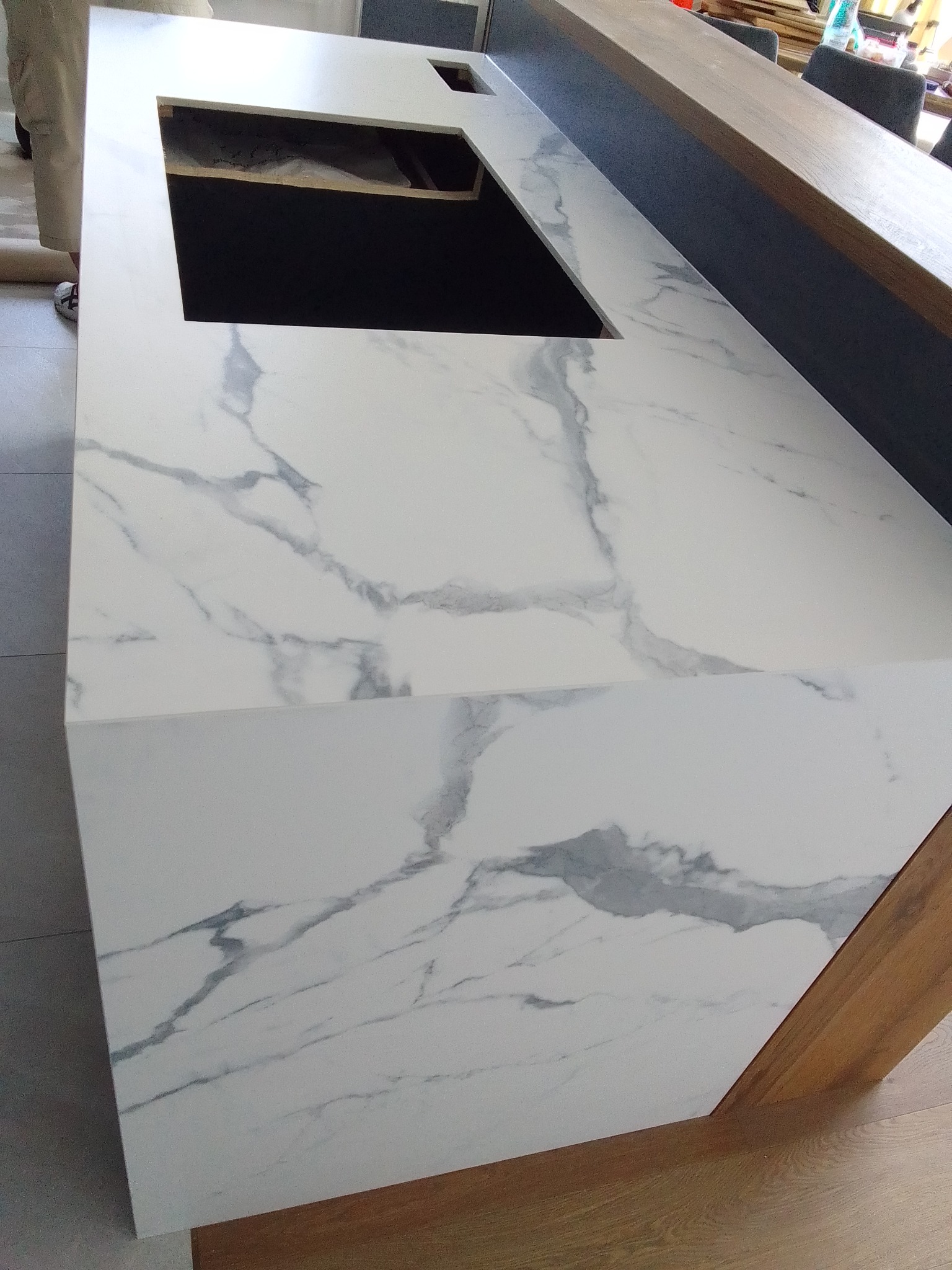 Biały blat z marmurowym wzorem, wycięty otwór na zlew, widoczny fragment nogi osoby w tle, montaż blatu w trakcie.