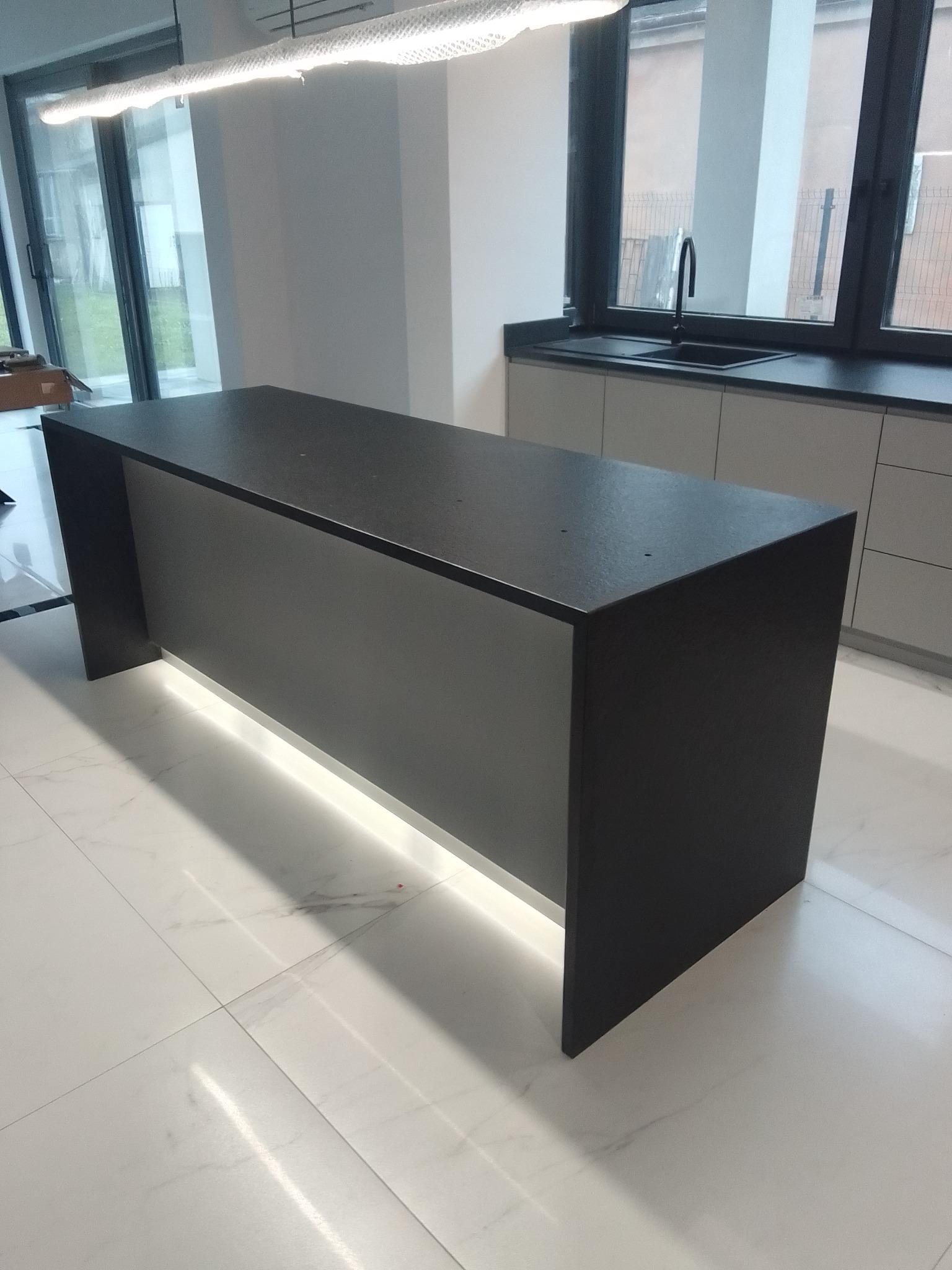 Czarny, matowy blat kuchenny z podświetleniem LED u dołu, na tle jasnej kuchni z oknem i zlewem. Minimalistyczny design, nowoczesne wykończenie.