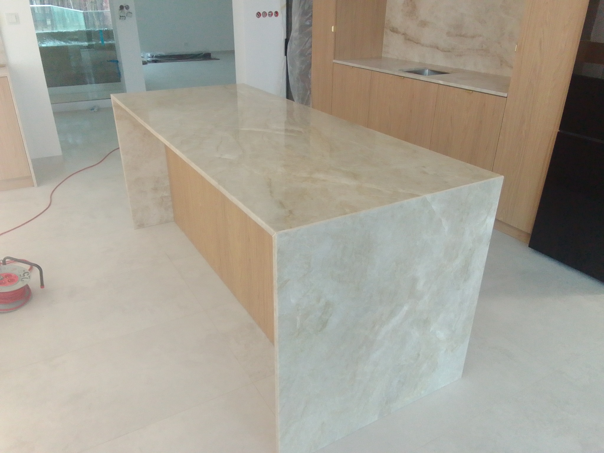 Beżowy blat kamienny z wyraźnym wzorem żyłkowania, tworzący wyspę kuchenną. Widoczne szafki w tle i jasna podłoga. Uwagę przykuwa połyskująca powierzchnia blatu.