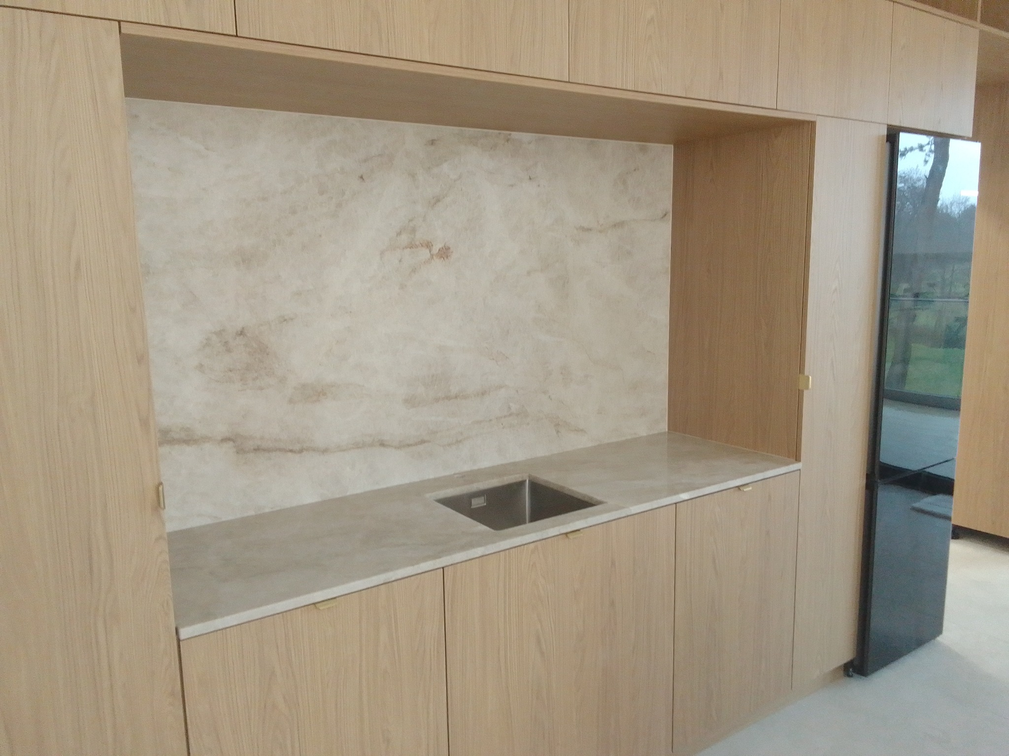 Jasna kuchnia na wymiar z blatem kamiennym i marmurowym panelem nad zlewem. Minimalistyczny design, drewniane fronty szafek, nowoczesne wykończenie.