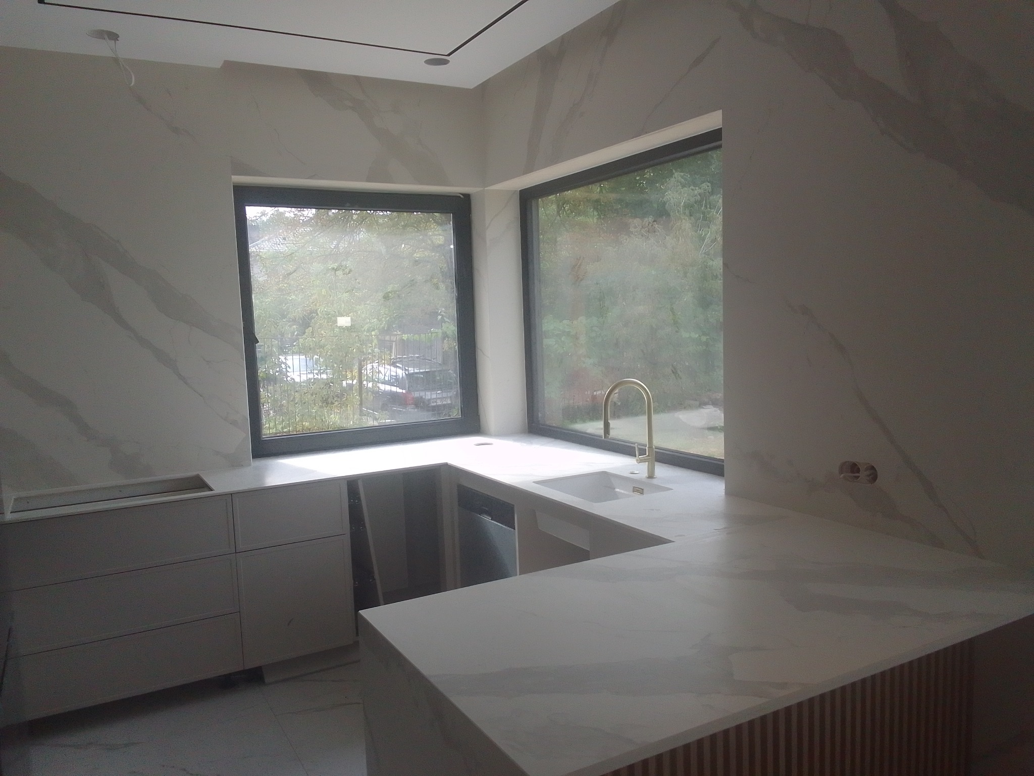 Nowoczesna kuchnia z blatem z kamienia w odcieniach bieli i szarości, złota bateria, duże okna z widokiem na zieleń. Minimalistyczny design, marmurkowy wzór na ścianach i blacie.