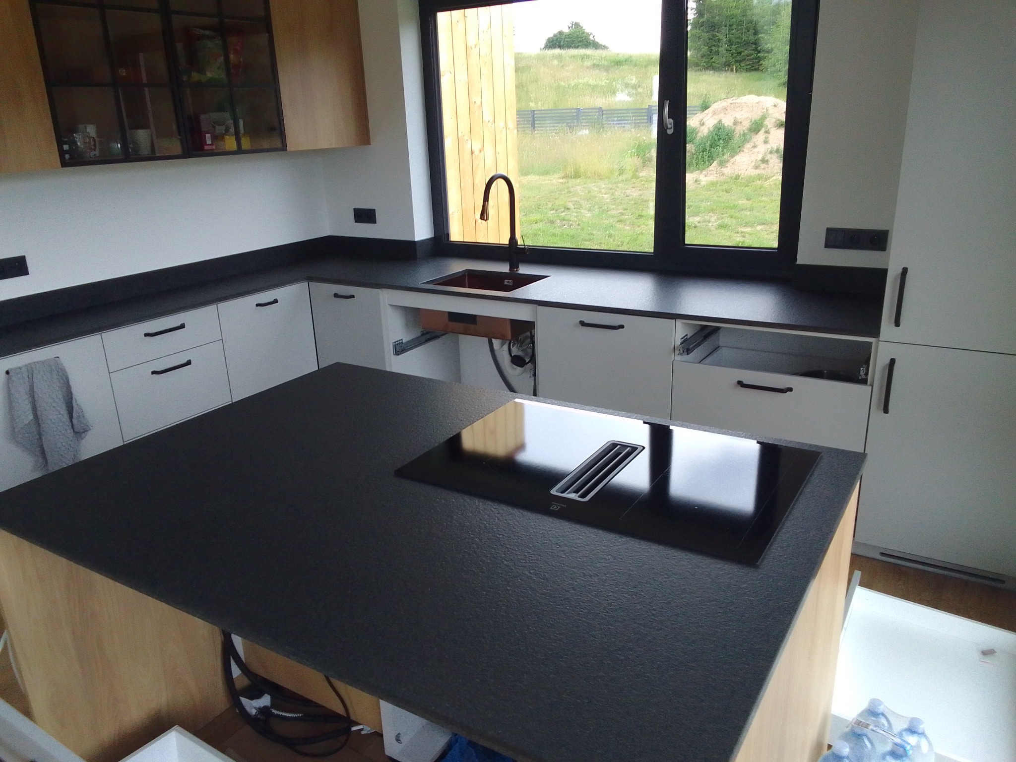 Nowoczesna kuchnia z wyspą i czarnym blatem kamiennym, minimalistyczne białe szafki, czarna armatura i widok na zieleń za oknem.