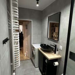 Marcin Florek Cactus Ink - Nowoczesna łazienka w Raciborzu z szarymi płytkami, czarną umywalką nablatową na drewnianym blacie, grzejnikiem drabinkowym i pralką, widoczna z perspektywy drzwi wejściowych.