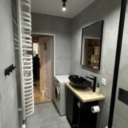 Nowoczesna łazienka w Raciborzu z szarymi płytkami, czarną umywalką nablatową na drewnianym blacie, grzejnikiem drabinkowym i pralką, widoczna z perspektywy drzwi wejściowych.