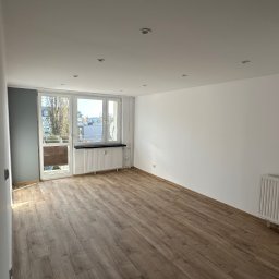 Marcin Florek Cactus Ink - Pusty pokój po odświeżeniu: nowe panele podłogowe w odcieniach brązu, świeżo pomalowane białe ściany z szarą ścianą akcentującą przy balkonie, widoczne grzejniki pod oknami.