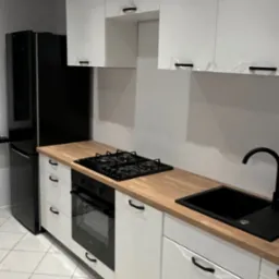 Nowoczesna kuchnia w bieli i czerni z drewnianym blatem, widoczne folie ochronne na frontach szafek sugerujące świeży montaż.