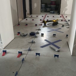 Marcin Florek Cactus Ink - Układanie jasnych płytek podłogowych z użyciem systemu poziomowania, widoczne narzędzia: poziomica, taśma, szlifierka kątowa. W tle surowe ściany w trakcie wykończenia.