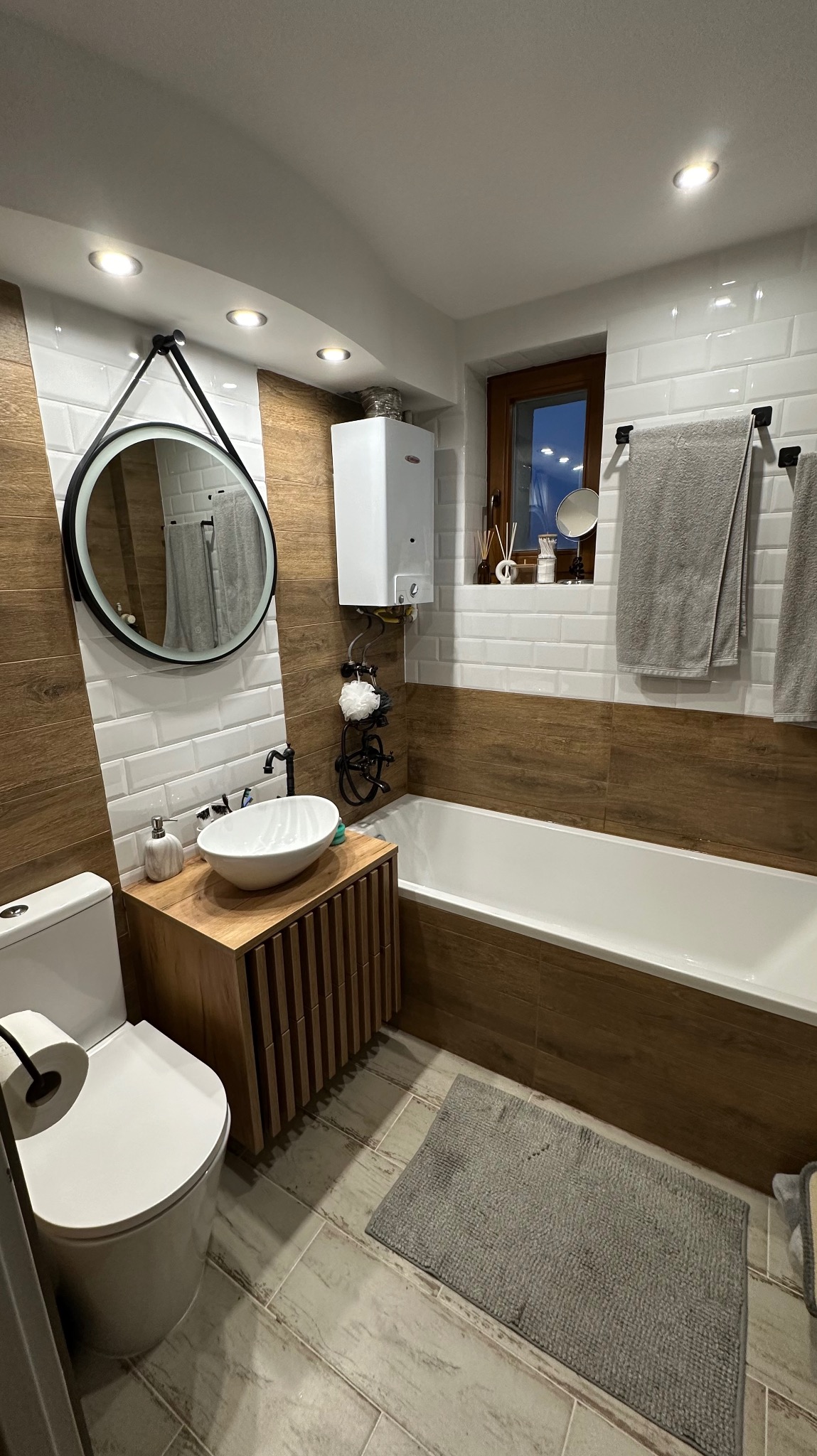 Nowoczesna łazienka po renowacji z drewnopodobnymi płytkami, okrągłym lustrem z podświetleniem, białą umywalką nablatową i grzejnikiem gazowym nad wanną.