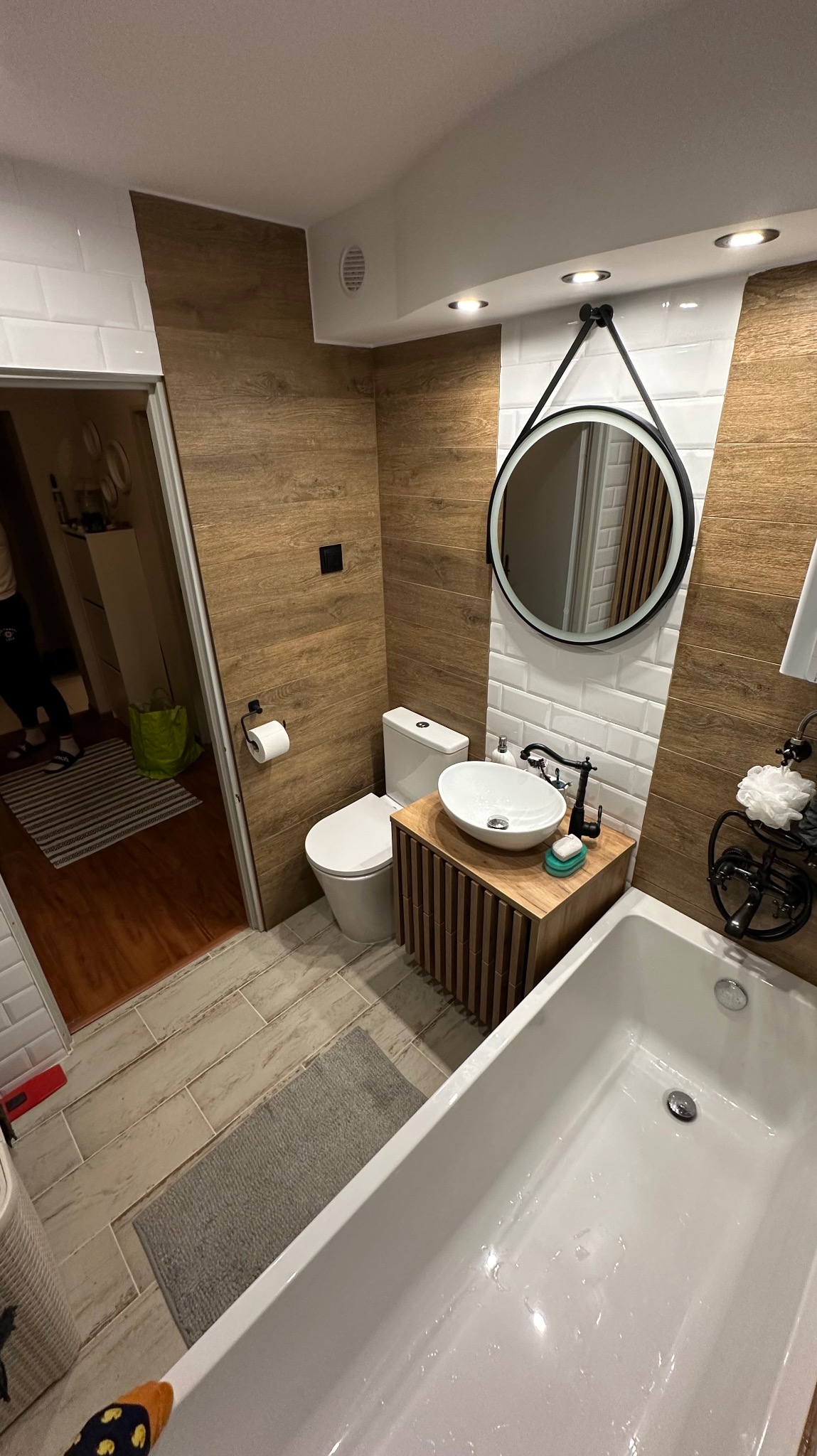 Nowoczesna łazienka z drewnianymi akcentami: ściana wyłożona panelami imitującymi drewno, okrągłe lustro z czarną ramą, biała umywalka nablatowa na szafce z lamelami, biała wanna oraz toaleta.