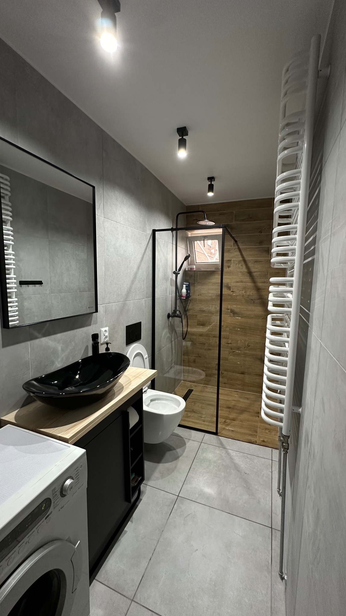 Nowoczesna łazienka w szarościach z czarnymi akcentami: umywalka nablatowa, kabina prysznicowa typu walk-in z drewnopodobnymi płytkami, biały grzejnik drabinkowy i pralka.