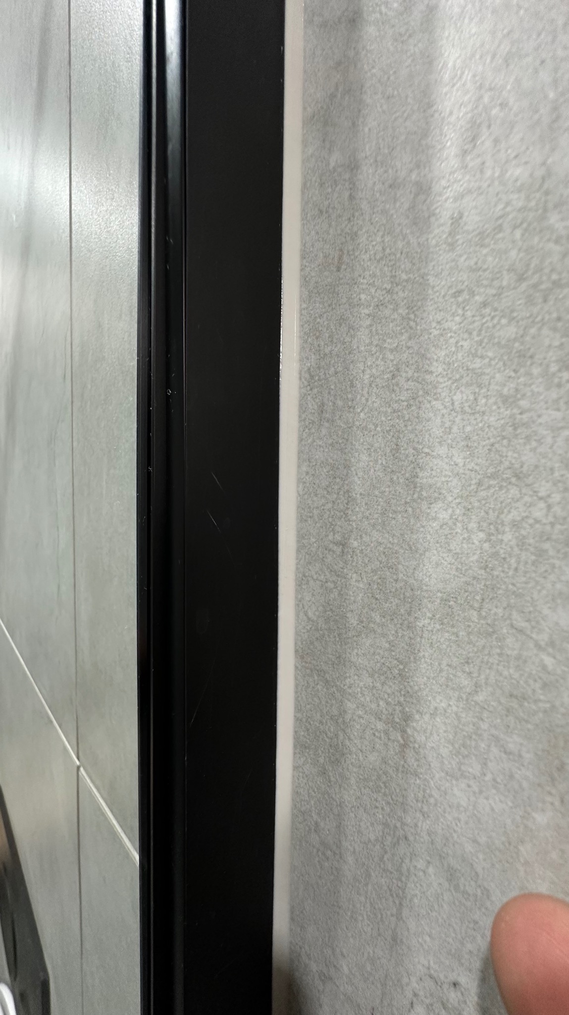 Fragment ściany z szarych płytek z czarną ramą, widoczny z bliska pod kątem, z fragmentem palca w prawym dolnym rogu.