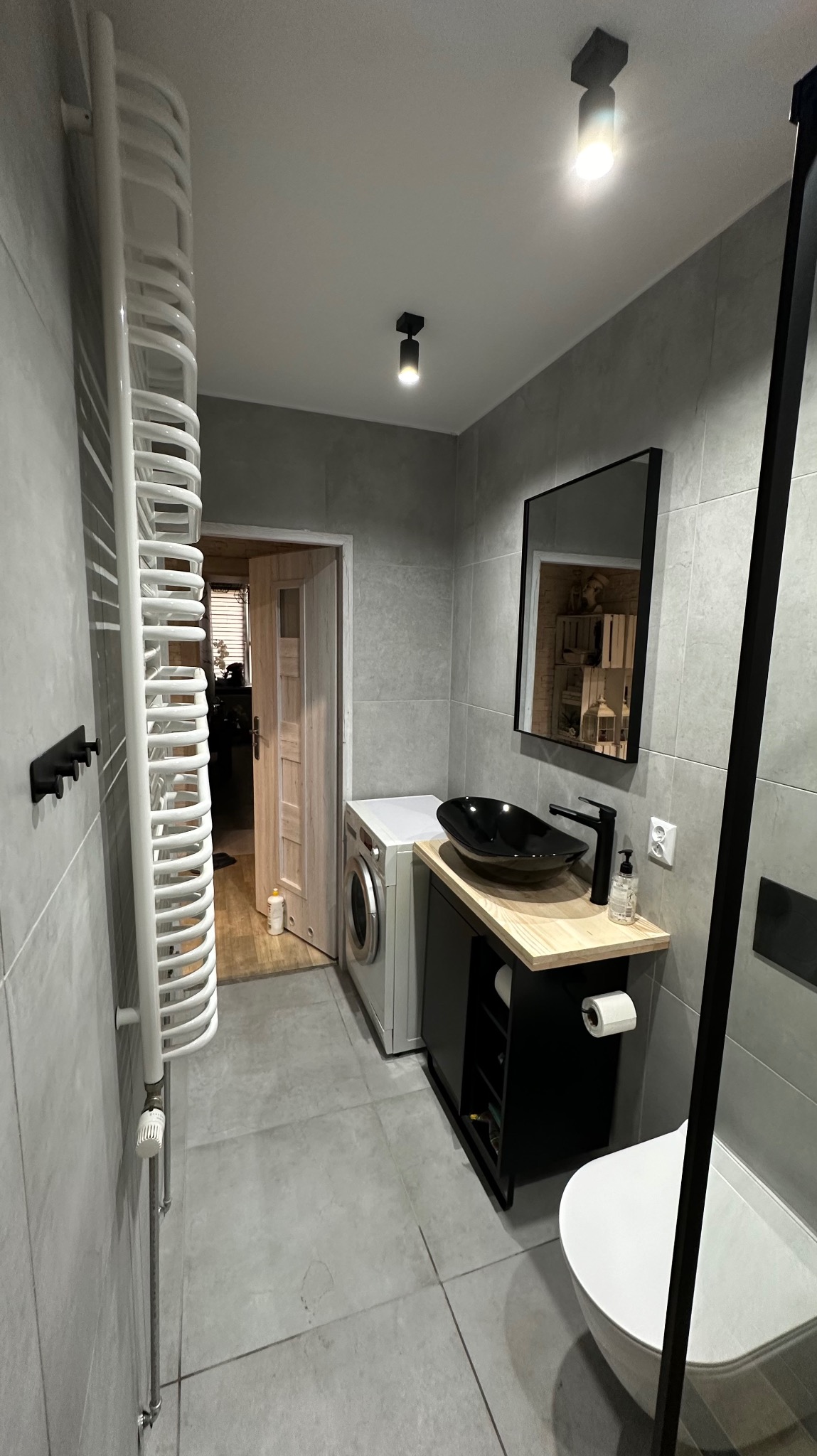 Nowoczesna łazienka w Raciborzu z szarymi płytkami, czarną umywalką nablatową na drewnianym blacie, grzejnikiem drabinkowym i pralką, widoczna z perspektywy drzwi wejściowych.