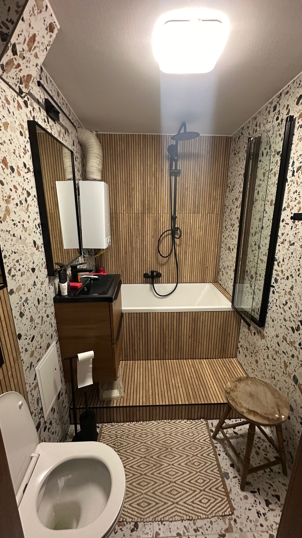 Wnętrze wyremontowanej łazienki z drewnianymi panelami na ścianie i podłodze, wzorzystą terakotą na pozostałych ścianach, czarną armaturą i drewnianym stołkiem.