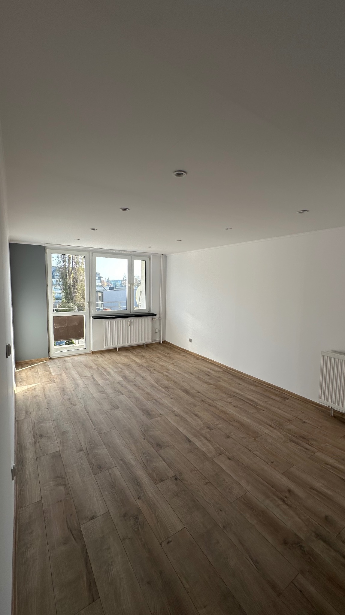 Pusty pokój po odświeżeniu: nowe panele podłogowe w odcieniach brązu, świeżo pomalowane białe ściany z szarą ścianą akcentującą przy balkonie, widoczne grzejniki pod oknami.