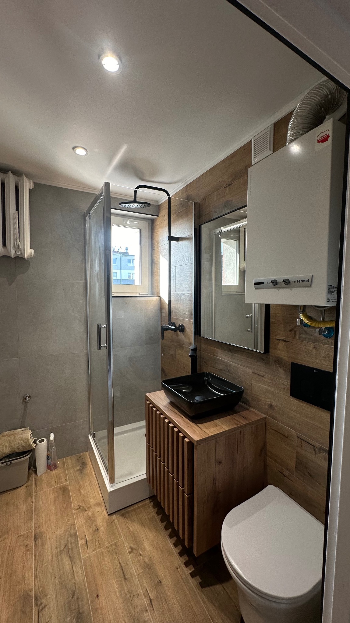 Nowoczesna łazienka z kabiną prysznicową, umywalką nablatową na drewnianej szafce, podwieszanym WC i grzejnikiem gazowym, wykończona płytkami imitującymi drewno i szarością betonu.