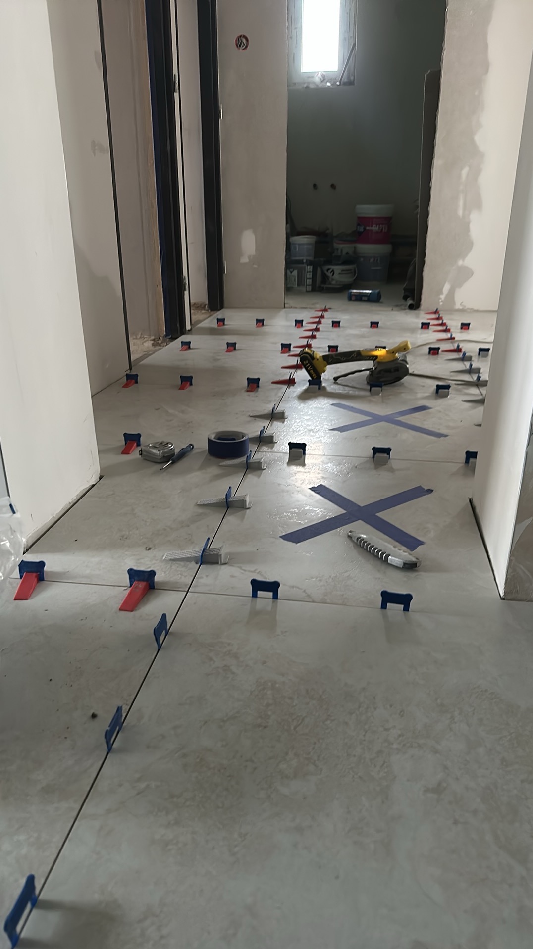 Układanie jasnych płytek podłogowych z użyciem systemu poziomowania, widoczne narzędzia: poziomica, taśma, szlifierka kątowa. W tle surowe ściany w trakcie wykończenia.