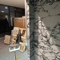 Serhii Horodetskyi - Wykończona ściana z płytek imitujących marmur, widoczna przez futrynę, w tle palety z materiałami budowlanymi i lampa budowlana.