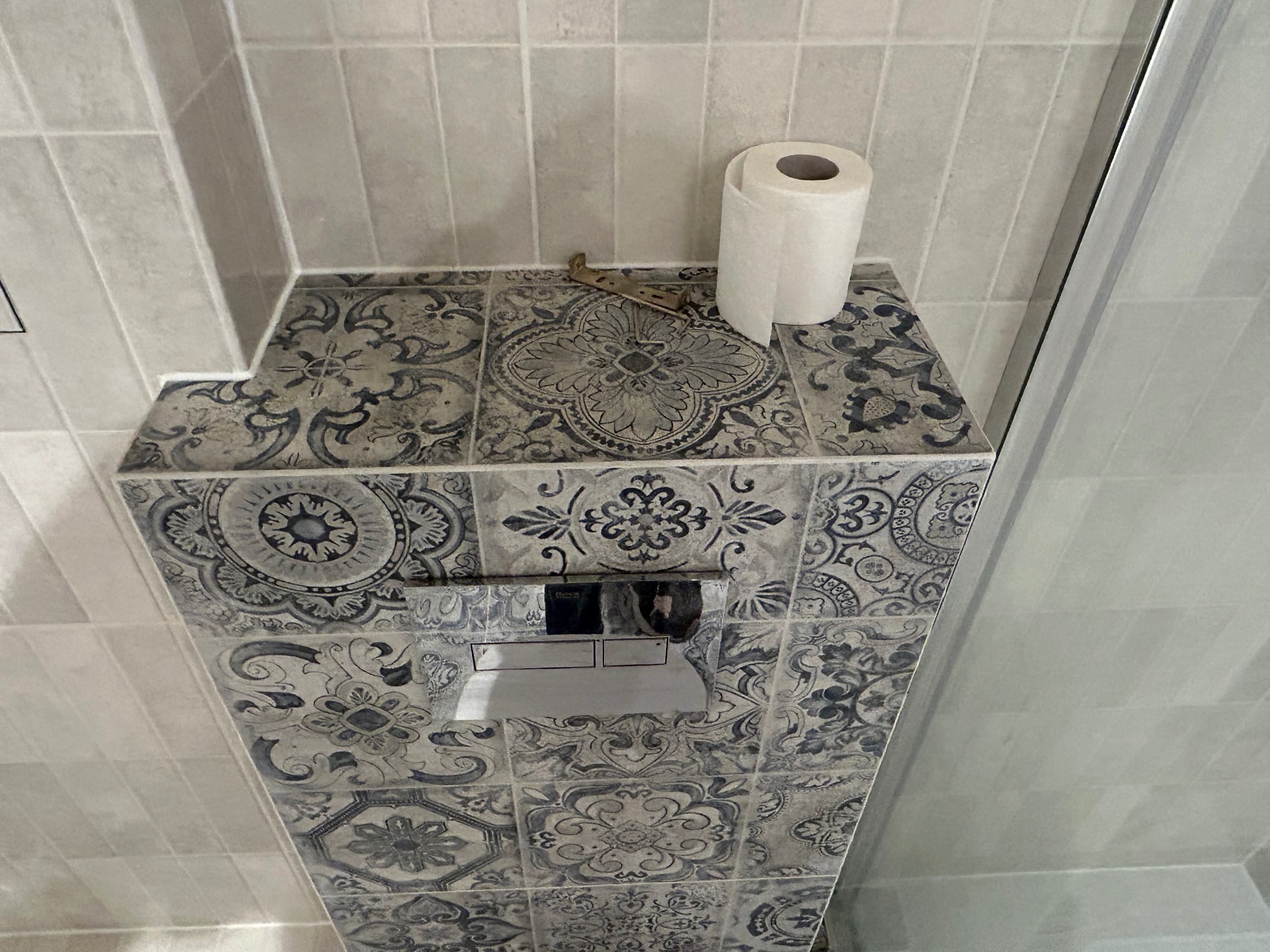 Obudowa stelaża WC z dekoracyjnymi płytkami w stylu vintage, rolka papieru toaletowego i metalowy element na górze, kremowe płytki na ścianie, fragment kabiny prysznicowej.