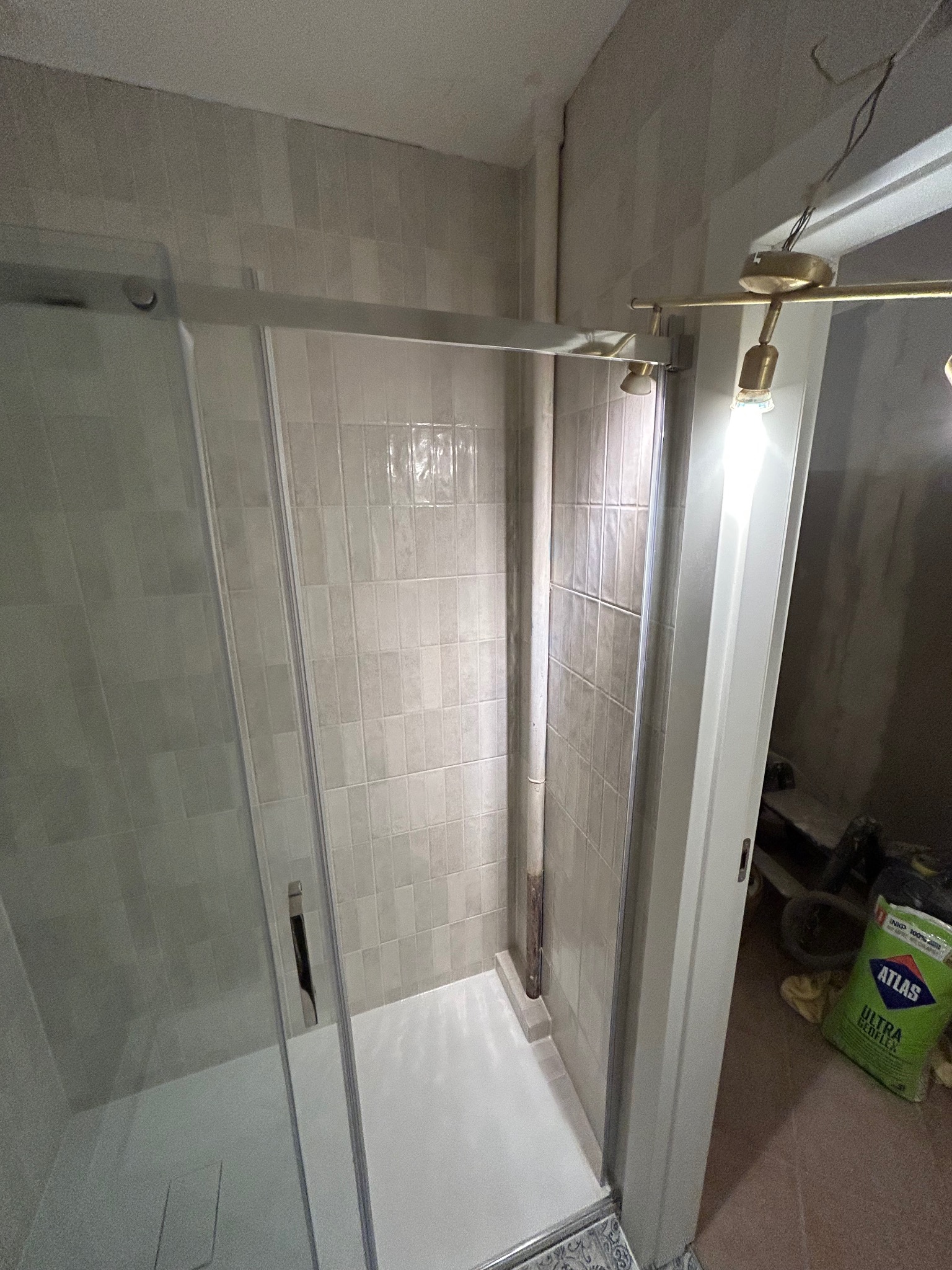 Zamontowana kabina prysznicowa z przesuwnymi drzwiami, wykończona beżowymi płytkami, widoczny worek z klejem Atlas Ultra Geoflex w tle, sugerujący prace wykończeniowe.