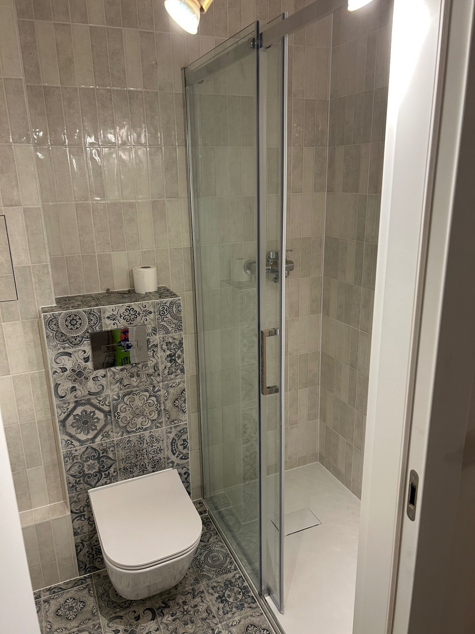 Wykończona łazienka z kabiną prysznicową z przesuwnymi drzwiami, wiszącą toaletą na tle ściany z dekoracyjnymi płytkami oraz beżowymi płytkami na ścianach kabiny.