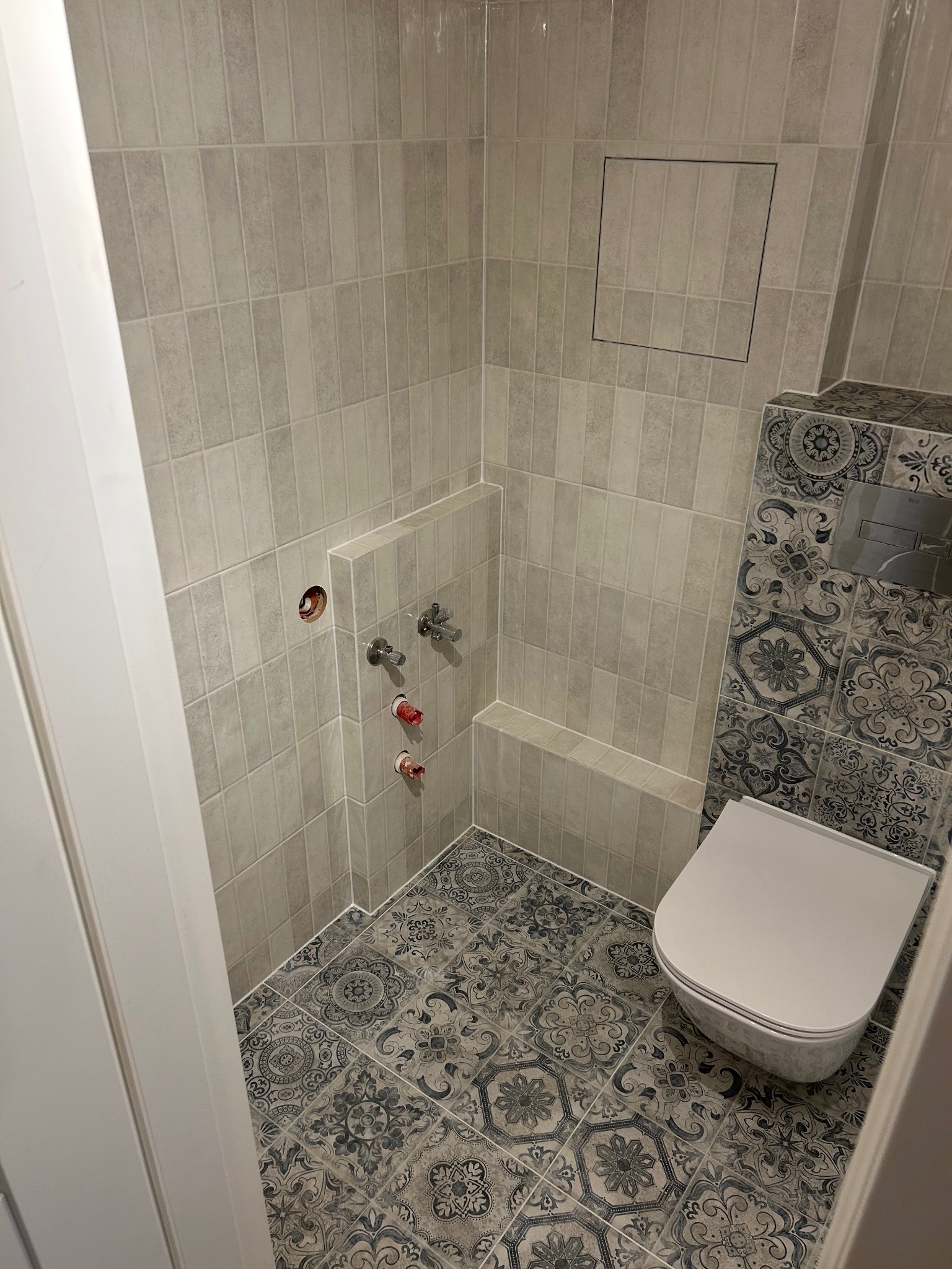 Wykończona łazienka z szaro-beżowymi, pionowo ułożonymi płytkami na ścianach, dekoracyjnymi płytkami z motywem kwiatowym przy toalecie i na podłodze. Widoczne wystające rury i zawory oraz otwór...