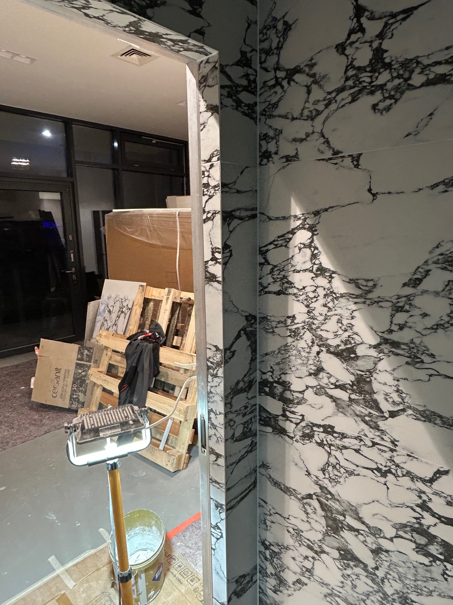 Wykończona ściana z płytek imitujących marmur, widoczna przez futrynę, w tle palety z materiałami budowlanymi i lampa budowlana.