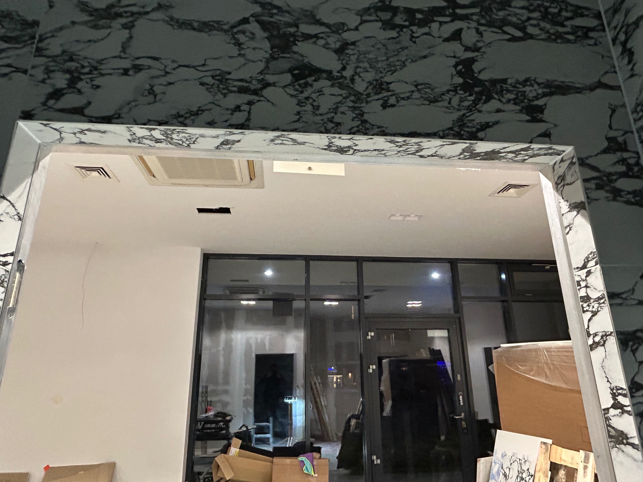 Wnętrze w trakcie prac wykończeniowych: widoczna marmurowa okładzina na ścianach i framudze, sufit z kratkami wentylacyjnymi oraz szklane drzwi z czarną ramą. W tle widoczne kartony i narzędzia...