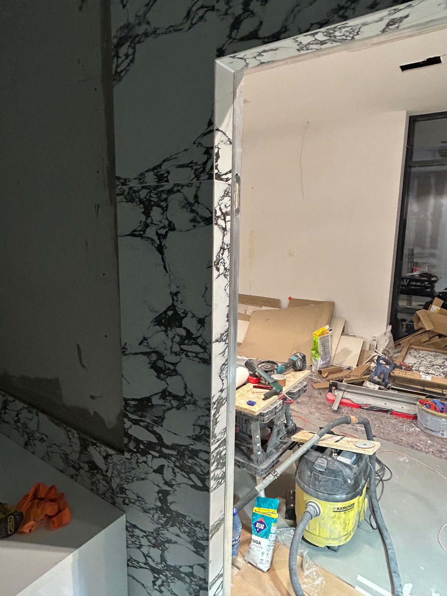 Fragment łazienki w trakcie prac wykończeniowych: ściana z płytek imitujących marmur, widoczny odkurzacz przemysłowy Karcher, narzędzia i materiały budowlane w tle, w tym fuga Atlas.