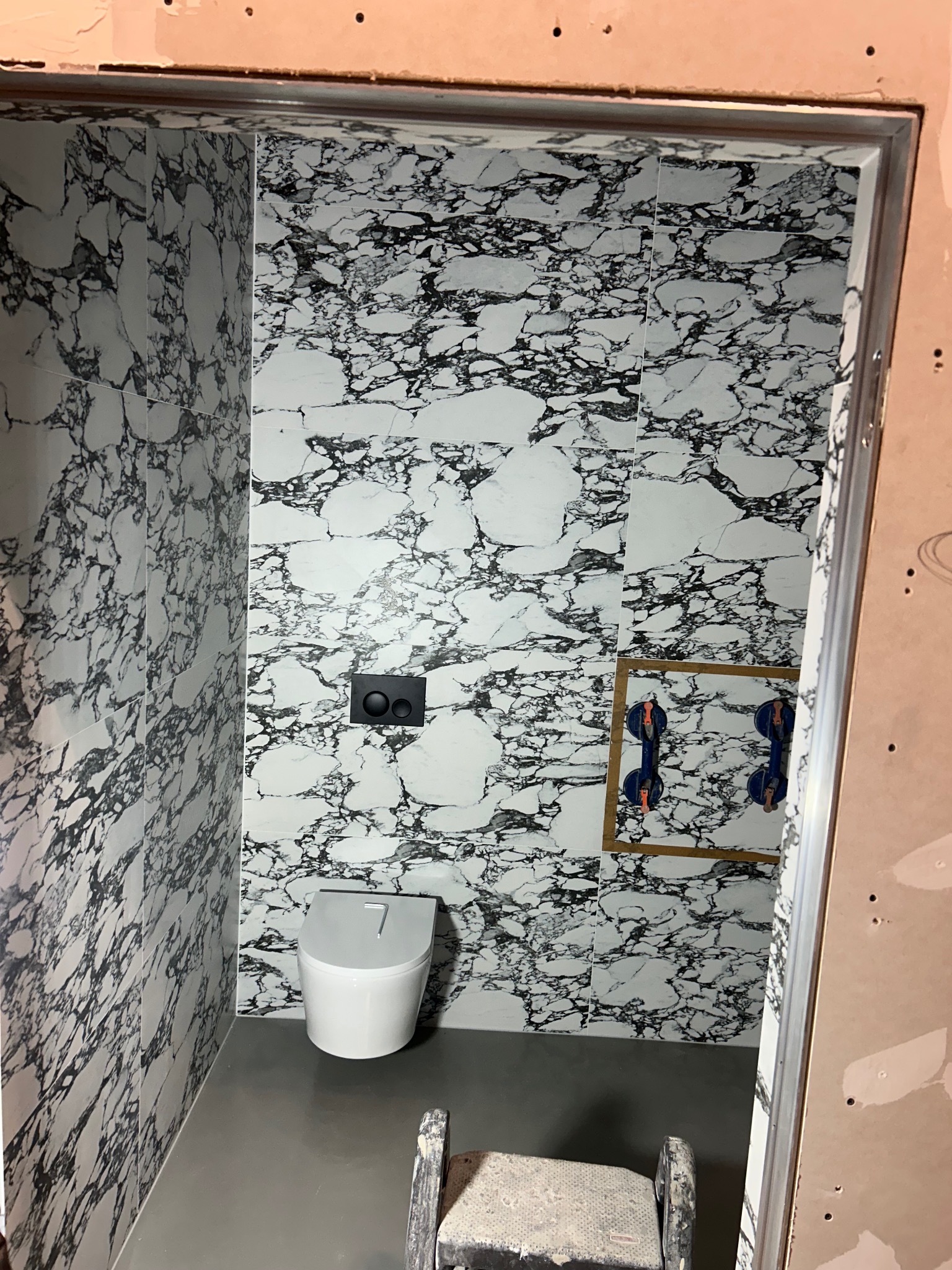 Wnętrze łazienki w trakcie remontu, ściany wyłożone płytkami imitującymi marmur, wisząca miska WC, widoczne przyssawki do płytek na ścianie, szary podkład na podłodze, fragment drabiny malarskiej.
