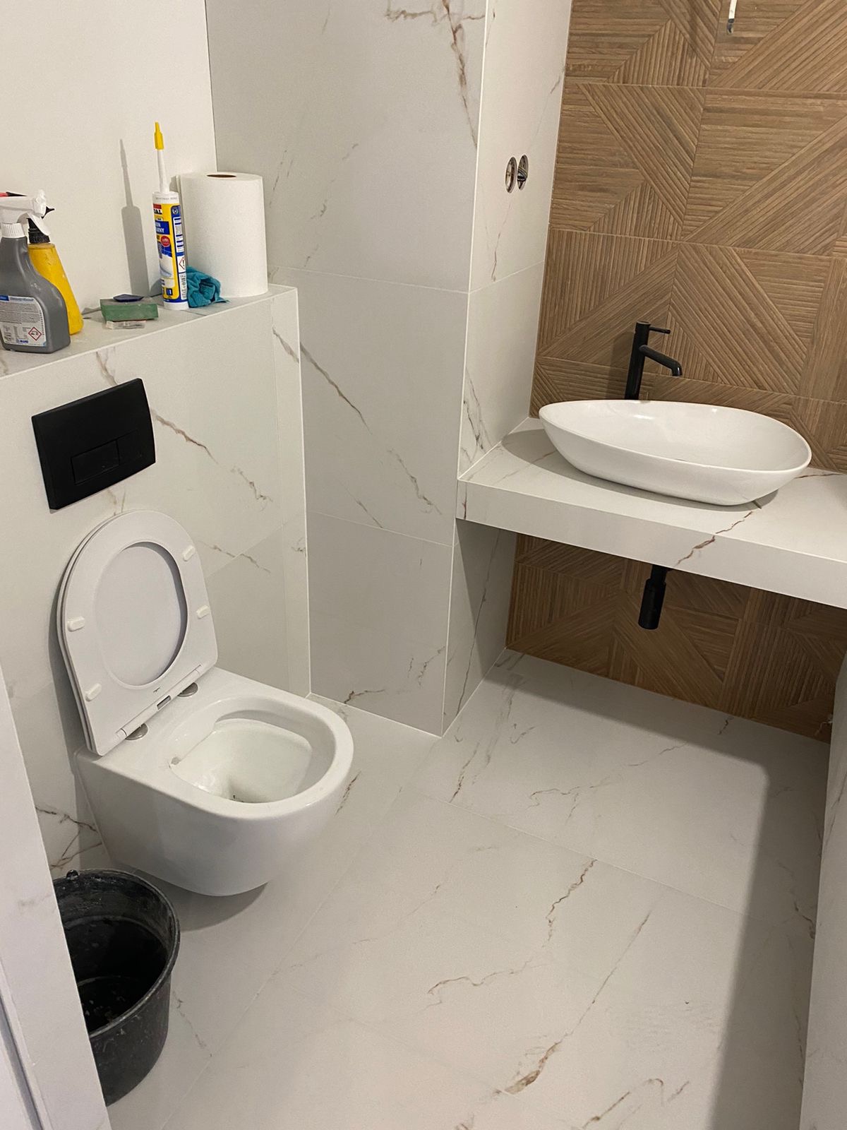 Nowoczesna łazienka z wiszącą toaletą, białą umywalką na blacie i dekoracyjnymi panelami ściennymi z drewna, widoczne akcesoria i wiadro, świeżo po remoncie.