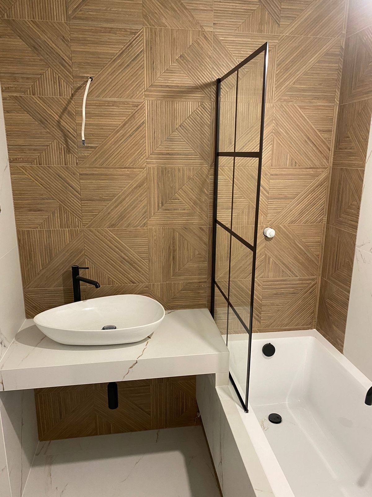 Nowoczesna łazienka z umywalką nablatową, kabiną prysznicową typu walk-in z czarnymi profilami i geometrycznymi płytkami imitującymi drewno na ścianie, widoczny wystający kabel.