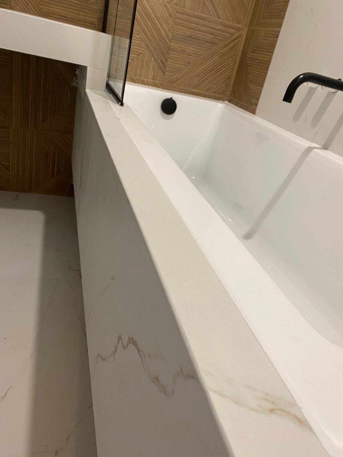 Biała wanna z zabudową z płytek imitujących marmur, czarna bateria, szklany panel prysznicowy i drewnopodobna ściana w łazience.