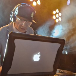 TOP 360 SPÓŁKA Z OGRANICZONĄ ODPOWIEDZIALNOŚCIĄ - DJ w szarej marynarce i czapce z daszkiem, grający na laptopie Apple, podświetlony przez reflektory i otoczony dymem scenicznym podczas występu.