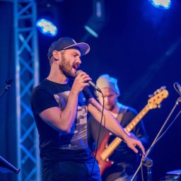 TOP 360 SPÓŁKA Z OGRANICZONĄ ODPOWIEDZIALNOŚCIĄ - Wokalista z brodą w czapce śpiewa do mikrofonu na scenie oświetlonej niebieskim światłem, w tle gitarzysta i perkusista.