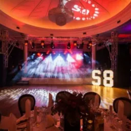 Sala balowa z oświetleniem scenicznym, dekoracjami świetlnymi w kształcie cyfr '58', stołami przygotowanymi do uroczystości i żyrandolem w tle.