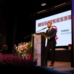 Prelegent w ciemnym garniturze przemawia na scenie podczas jubileuszu, za nim ekran z napisem 'Jubileusz' i hasłem 'Tradycja, która inspiruje', a po lewej stronie orkiestra smyczkowa.