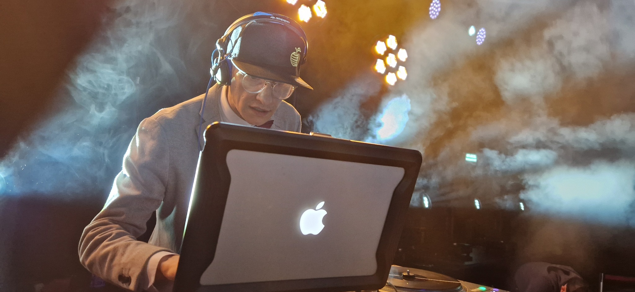 DJ w szarej marynarce i czapce z daszkiem, grający na laptopie Apple, podświetlony przez reflektory i otoczony dymem scenicznym podczas występu.