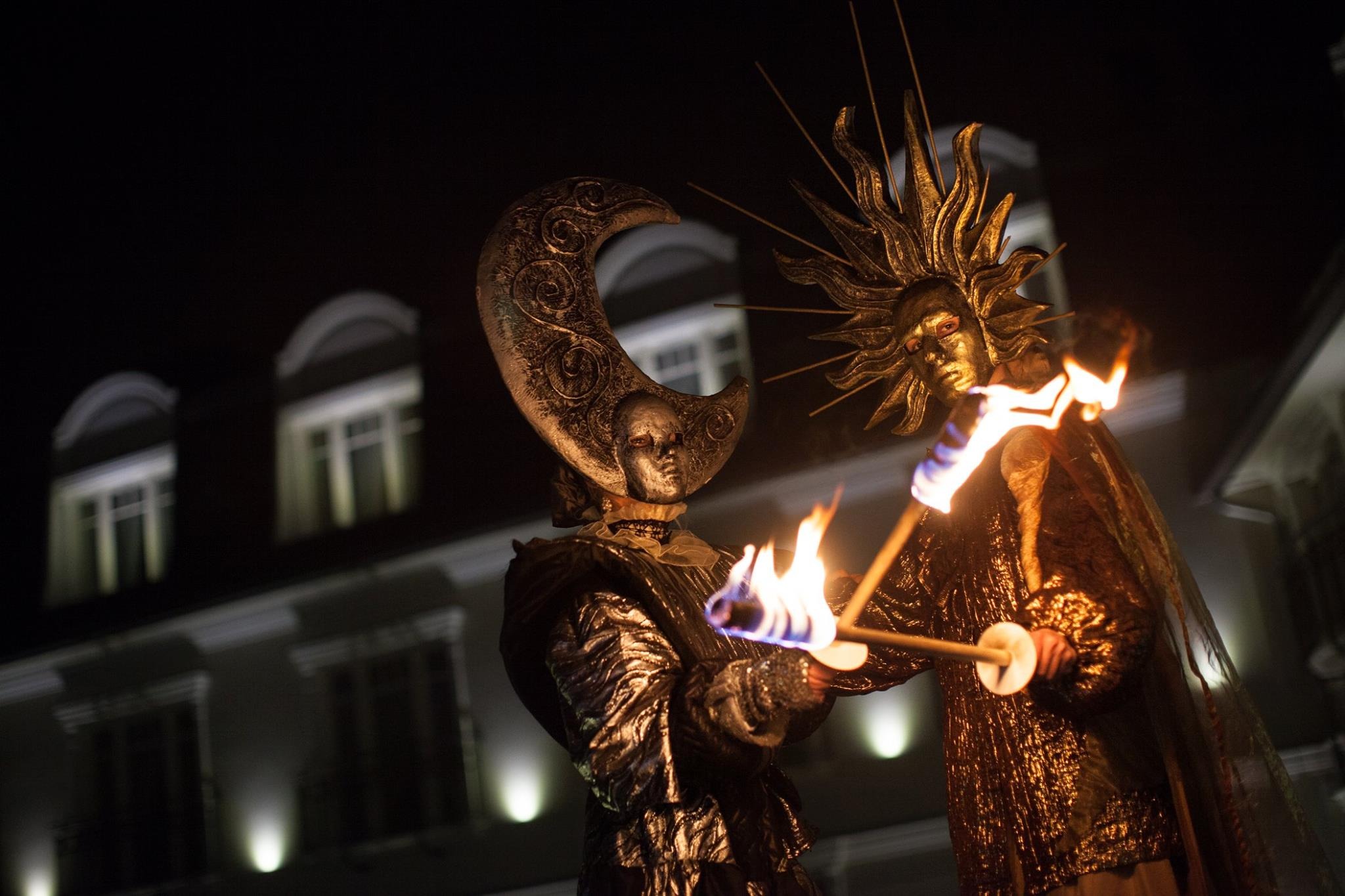 Dwie osoby w kostiumach Słońca i Księżyca z płonącymi pochodniami na tle budynku podczas nocnego pokazu.