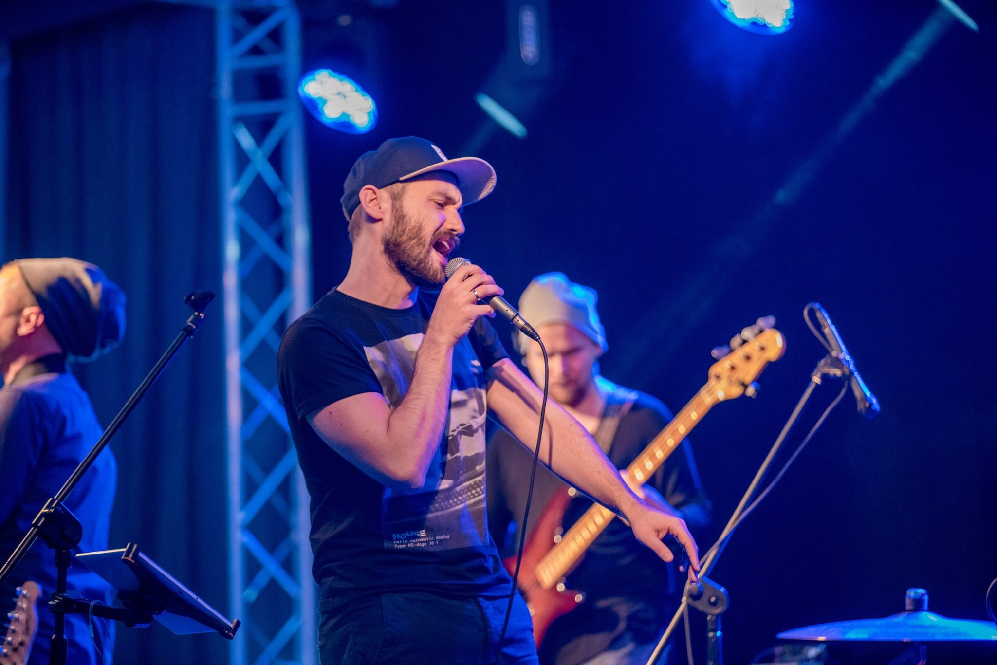 Wokalista z brodą w czapce śpiewa do mikrofonu na scenie oświetlonej niebieskim światłem, w tle gitarzysta i perkusista.