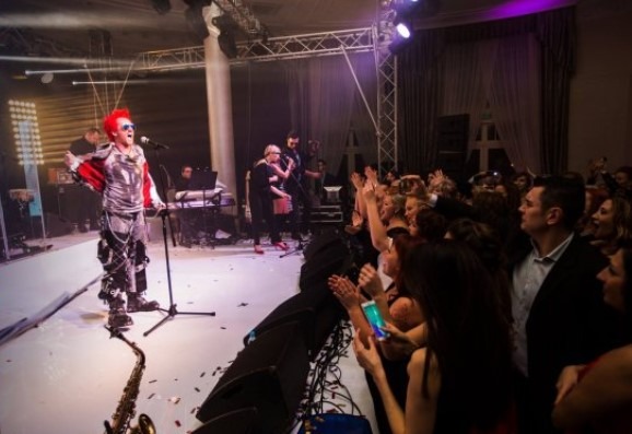 Występ zespołu muzycznego na scenie podczas imprezy, widownia klaszcze, oświetlenie sceniczne, saksofon na pierwszym planie.