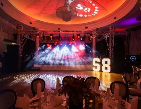 Sala balowa z oświetleniem scenicznym, dekoracjami świetlnymi w kształcie cyfr '58', stołami przygotowanymi do uroczystości i żyrandolem w tle.